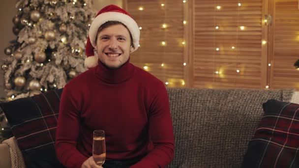 Porträt eines glücklich lächelnden kaukasischen Mannes mit Weihnachtsmütze und Winterpullover, der auf einer Couch in der Nähe des Weihnachtsbaums sitzt und ein Glas Champagner anhebt.. — Stockvideo