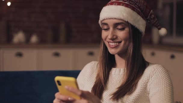 Красива усміхнена молода дівчина в капелюсі Сантас сидить на дивані біля різдвяної ялинки Прокрутка на її концепції смартфона відпустки та Новий рік крупним планом . — стокове відео