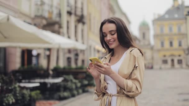 Atractiva chica joven con estilo usando su Smartphone escribiendo y sonriendo de pie en el fondo de la Ciudad Vieja . — Vídeos de Stock