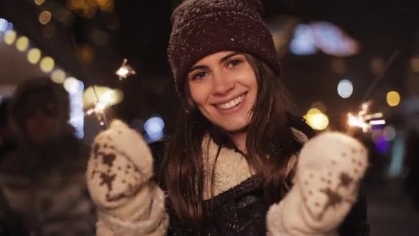 Acercamiento de Niza Chica Sonriente en Ropa de Invierno, Mirando Felizmente a la Cámara, Sosteniendo Chispas, De pie en la Caída de la Nieve en Navidad Feria Luces Fondo. Concepto de vacaciones . — Vídeo de stock