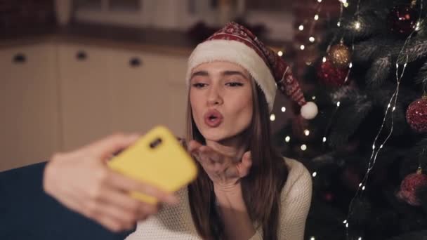 Glimlachend jong aantrekkelijk meisje dragen Santas hoed zittend op de Bank in de buurt van kerstboom maken Selfie poseren waait een kus en het gebruik van haar telefoon concept van vakantie en Nieuwjaar. — Stockvideo