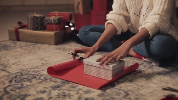 Close Up delle mani di Mulatto Girl Preparazione regali, Taglio carta da imballaggio, con regalo su di esso, Seduto sotto l'albero di Natale a casa accogliente sfondo. Concetto di celebrazione delle vacanze . — Video Stock