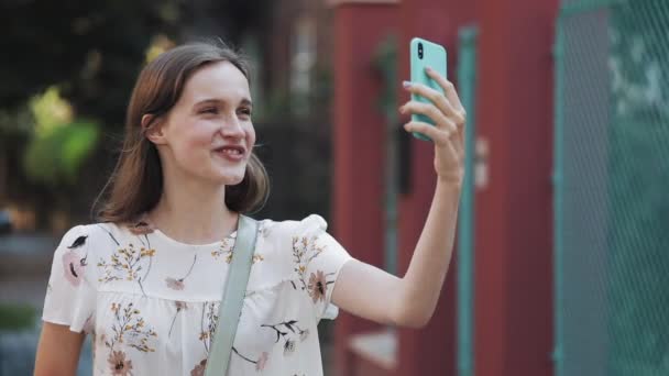 Pretty Caucasian Brown Hair Girl Wearing White Flower Dress with Bag Cross her Shoulder Using Her Smartphone and Making Video Message or Video Call Walking near Turquoise Fence Smiling. — Stock Video