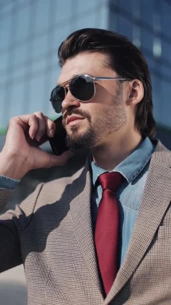 Вертикальное видео. Молодой привлекательный бизнесмен, выглядящий довольным, разговаривая по телефону, настраивает солнцезащитные очки, покачивая головой возле Центра Бизнеса . — стоковое видео