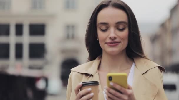 魅力的な若い女性は、彼女のスマートフォンを使用して笑顔のコフカップを保持し、シティストリートで歩いてタイピングクローズアップ. — ストック動画