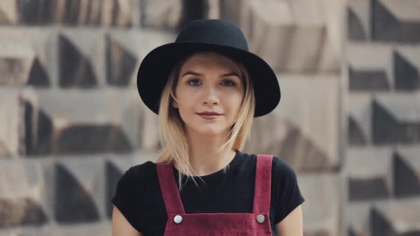 Портрет молодої білявою дівчиною з пронизливим в носі носіння чорної капелюсі повороту до камери і посміхаючись стоячи на міській вулиці — стокове відео
