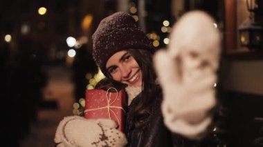 Gülümseyen Genç Charming Kız Yakın Portresi, Mutlu Görünümlü, Hediye Kutusu Holding, Düşen Kar Açık Ayakta, Noel Dekore Sokak Arka Planda kameraya el sallayarak. Tatil Konsepti.