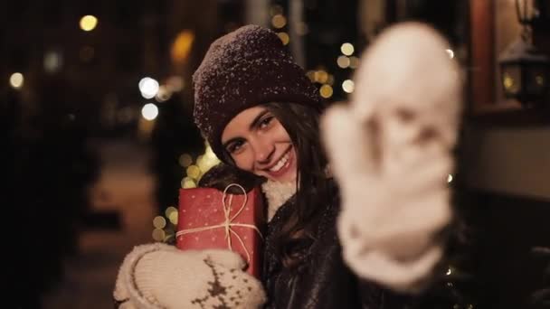 Primer plano Retrato de una joven y encantadora niña sonriente, luciendo feliz, sosteniendo la caja de regalo, de pie en la caída de la nieve al aire libre, saludando a la cámara en Navidad decorado fondo de la calle. Concepto de vacaciones . — Vídeo de stock