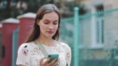 Yakın Çekim Beyaz Kahverengi Saç Kız Çanta Ile Beyaz Çiçek Elbise Giyen Onun Smartphone Ses Komutanı Kullanarak Omzuna Çapraz, Turkuaz Çit Gülümseme yakınında Yürüyüş Sesli Mesaj yapma.