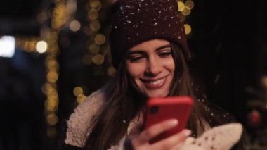 Kışın Gülümseyen Sevimli Kız Close Up Sosyal Ağlarda Sohbet, Onun Smartphone kullanarak, Ekrana Bakarak, Noel Dekore Akşam Karlı sokakta Yürüyüş.