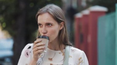 Kahverengi saçlı Mavi Gözlü ve Çiller ilerlik Beyaz Elbise Giyen Güzel Genç Gülümseyen Kafkas Kız Çiçeklerle Coffe Kupası Tutan ve Sokakta Yürüyüş İçme. Yakın Çekim.