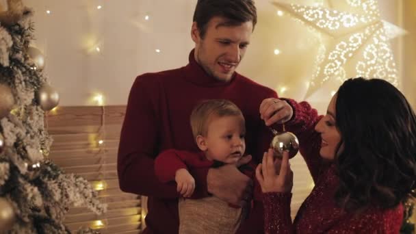 特写 年轻 美丽的 母亲 英俊 的父亲 和 可爱的 婴儿 儿子 在 父亲 手臂 附近的 圣诞树 微笑， 母亲显示男孩一个圣诞球装饰概念的家庭假期和新年. — 图库视频影像