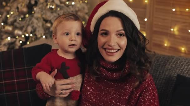 Nahaufnahme einer lächelnden schönen Mutter mit Weihnachtsmütze, die ihren kleinen Jungen auf dem Sofa im Wohnzimmer in der Nähe des Weihnachtsbaums hält, der Geschenke entgegennimmt und zurückgibt.. — Stockvideo