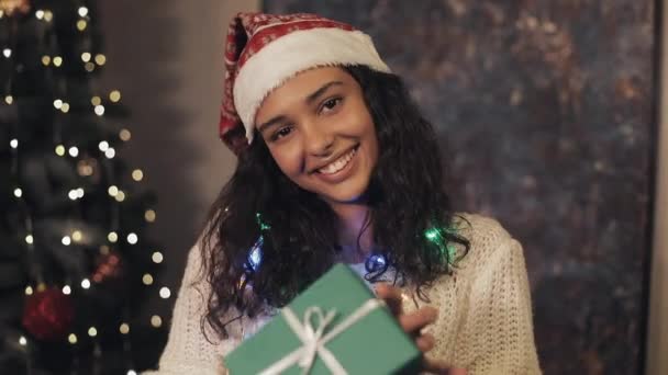 Jonge mooie mulatto vrouw bij Santas hoed en decoratieve lichten op haar nek houden aanwezig vak in haar handen terwijl staan in de buurt van kerstboom op gezellige thuis achtergrond. Vakantie viering concept. — Stockvideo