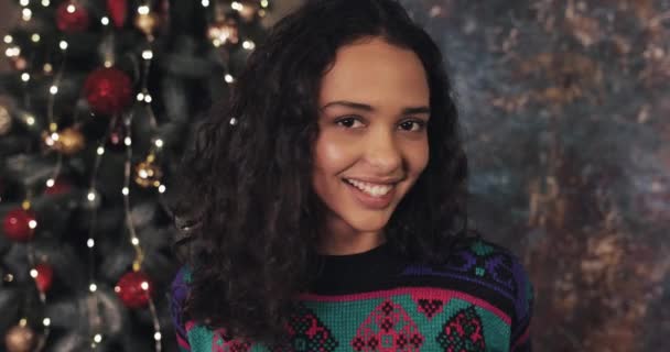 Portret van aantrekkelijke jonge Brunnete vrouw met een stralende glimlach, staande in de buurt van kerstboom, glimlachend en op zoek naar camera. Kerst en Nieuwjaar viering concept. — Stockvideo