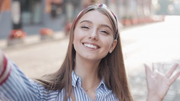 Menina atraente nova vestindo em camisa listrada com headband acenando Olá no bate-papo de vídeo Conversação Selfie com Smartphone e mostrando Okey sorrindo em pé na rua da cidade . — Vídeo de Stock