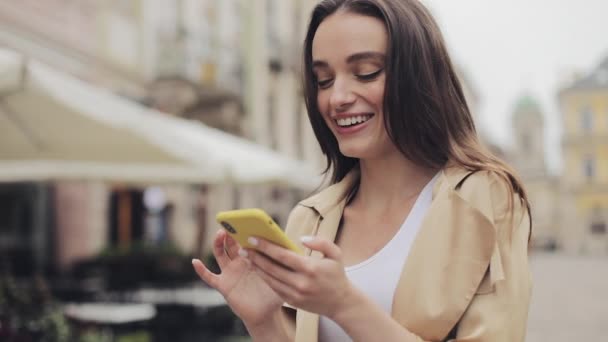 Приваблива стильна молода дівчина використовує свій смартфон друкуючи і посміхаючись стоячи на тлі Старого міста крупним планом . — стокове відео