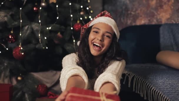 サンタの帽子と白いプルオーバーを身に着けた満足のいく若い女の子のクローズアップ, カメラの笑顔に赤いギフトボックスを伸ばす, 満足して見て, クリスマスの装飾された家に座って.ホリデーコンセプト. — ストック動画
