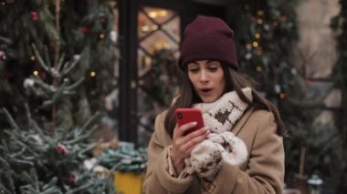 Pretty Girl Kış Şapka ve Eldivenler Onun Smartphone kullanarak sohbet, Ekrana Bakarak, Şaşırmış Arıyorsunuz ve Wow Ayakta Açık Dekore Noel Vitrini de Söyleyerek.