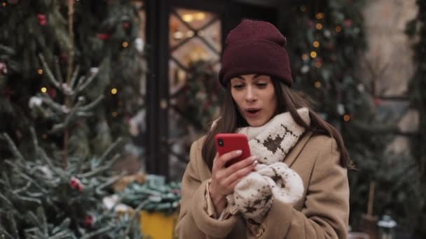 漂亮的女孩在冬季帽子和米滕斯聊天使用她的智能手机，看着屏幕，看着惊讶和说哇站在户外装饰圣诞窗店. — 图库视频影像