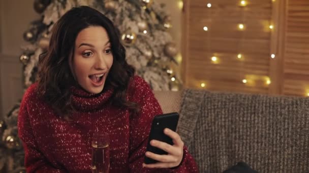 Portret van verbaasd jonge mooie vrouw het houden van een glas Champagne met behulp van haar Smartphone zitten in de buurt van kerstboom concept van vakantie en Nieuwjaar. — Stockvideo