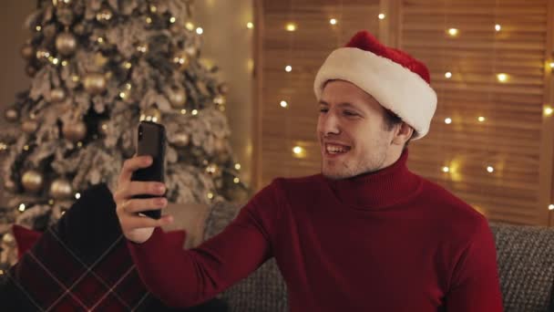 Homem caucasiano atrativo alegre que desgasta o chapéu de santas que Siiting no sofá perto da árvore de Natal usando seu smartphone para fazer uma chamada video ou um conceito video da mensagem dos feriados e da opinião lateral do ano novo. — Vídeo de Stock