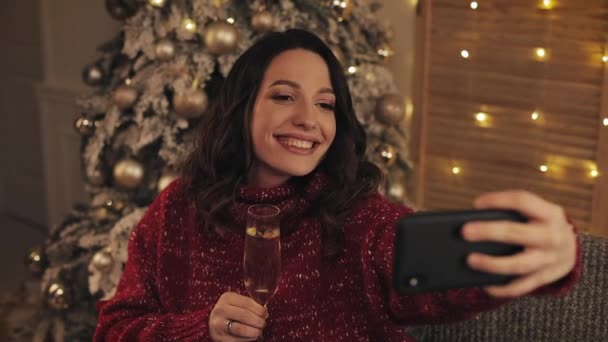 Gyönyörű fiatal barna nők viselése téli pulóver ülő közelében karácsonyfa otthon háttér Holding pezsgő üveg pózol készítése Selfies fogalma ünnepek és újév. — Stock videók