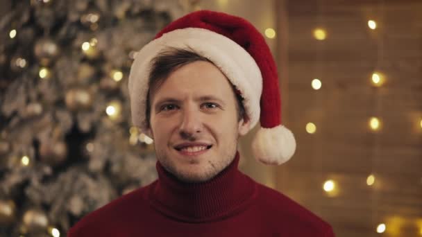 Indoor Close Up Portrait of Handsome Smiling Man in Santas Hat Sitting at Sofa near Christmas Tree and Sparkling Lights Background Concept of Holidays and New Year. — Stock Video