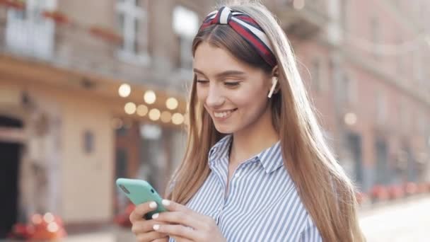 Menina bonita nova com cabelos castanhos e headband elegante vestindo camisa listrada e fones de ouvido usando Smartphone Sorrindo e olhando para câmera andando na rua da cidade . — Vídeo de Stock