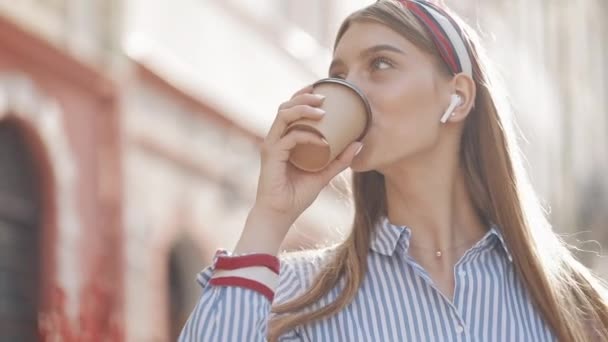 Całkiem zadowolony młoda kaukaski dziewczyna z brązowymi włosami i pałąka w paski koszula w słuchawkach uśmiechnięta picie kawy z Paper Cup chodzenie na ulicy City Street. Zbliżenie. — Wideo stockowe