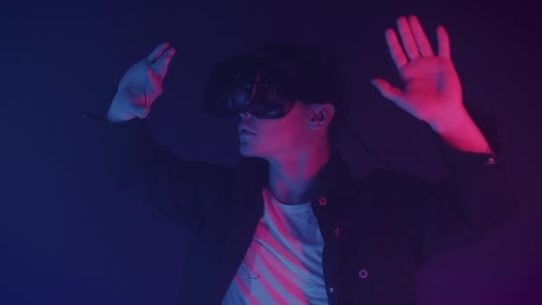 Aantrekkelijke man in virtual reality bril spelen spel en bewegende handen in de lucht op zoek onder de indruk, zeggen wow staande in de kamer met neon verlichting kleuren Futurictic achtergrond. — Stockvideo