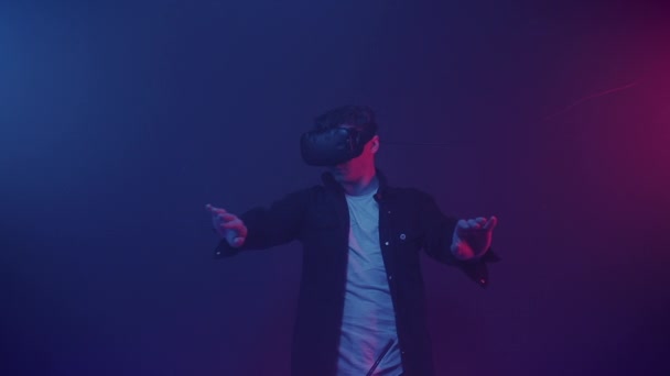 Hombre joven caucásico con auriculares de realidad virtual, juego, manos en movimiento en el aire, mecanografía, de pie en la habitación con fondo abstracto de colores de iluminación de neón. Concepto futuro . — Vídeos de Stock