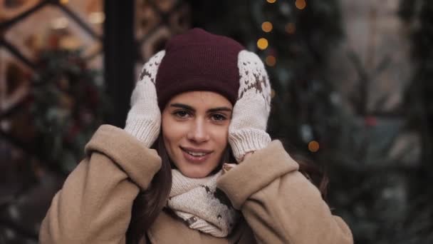 Zbliżenie Portret młodej uroczej dziewczyny na sobie szalik, rękawice, wprowadzenie jej kapelusz prosto i uśmiechnięty, patrząc do aparatu, stojący na Boże Narodzenie zdobione okno sklep tło. — Wideo stockowe