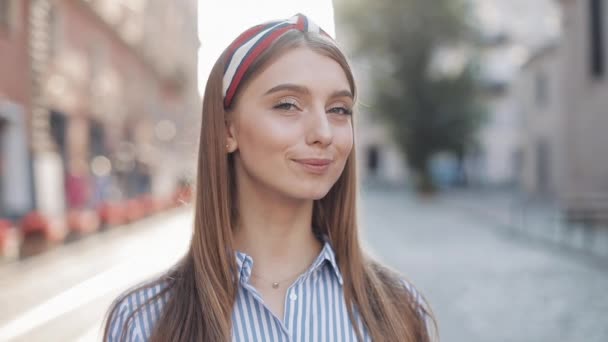 Portret zbliżenie młodych szczęśliwy dziewczyna z brązowe włosy i niebieskie oczy noszenie w paski sukienka i modny pałąk uśmiechnięty patrząc na aparat stojący na ulicy City. — Wideo stockowe