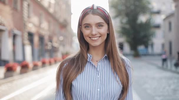 Portret van jong gelukkig charmant meisje met rechte bruin haar en blauwe ogen dragen in gestreepte jurk en modieuze hoofdband glimlachend op zoek naar camera staande op de City Street zoom. — Stockvideo