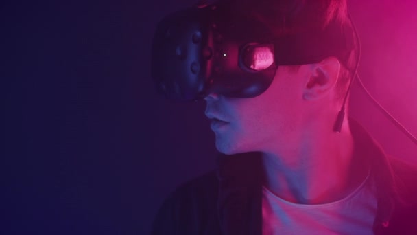 Fermer Portrait d'un homme portant un casque de réalité virtuelle levant les yeux et souriant debout dans la pièce avec un fond abstrait de couleurs néon. Concept futur . — Video