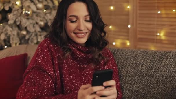 Ritratto di attraente giovane donna bruna che utilizza il suo smartphone digitando seduta vicino all'albero di Natale al divano grigio concetto interno di vacanze e capodanno . — Video Stock