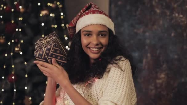 年轻的漂亮穆拉托女人在圣诞老人帽子和装饰灯在她的脖子上摇着现在盒子在她的手中，看着相机和微笑在舒适的家庭背景。节日庆典概念. — 图库视频影像