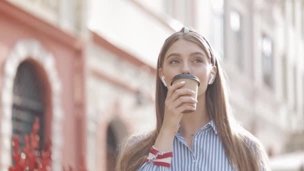 Портрет веселий усміхнений молодий красива дівчина носить стильний заставку і смугаста сорочка в навушники, пити каву, ходьба щасливо і озираючись на старому тлі міста. — стокове відео
