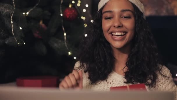 Närbild av ung vacker kvinna i Santas hatt med videochatt på sin bärbara dator hemma nära julgran, visar julklapp Box till kamera. Nytt år och julfirande koncept. — Stockvideo