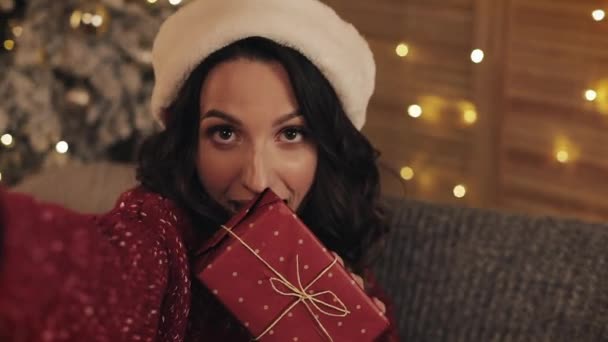 Atrakcyjne młoda brunetka kobiety z Santas Hat siedzi na kanapie w pobliżu choinki w przytulnym domu tle gospodarstwa Gift Box stwarzające Making selfie — Wideo stockowe