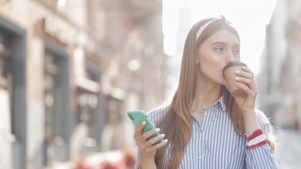 Retrato de la atractiva chica feliz caucásica con diadema usando su teléfono inteligente y sonriendo bebiendo café caminando en el fondo de la ciudad vieja de cerca. Concepto de comunicación . — Vídeo de stock