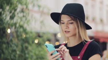 Akıllı telefonuna siyah şapkalı ve ayakta duran güzel sarışın kız şehrin arka planında kameraya bakmaktansa siyah şapkalı kız..