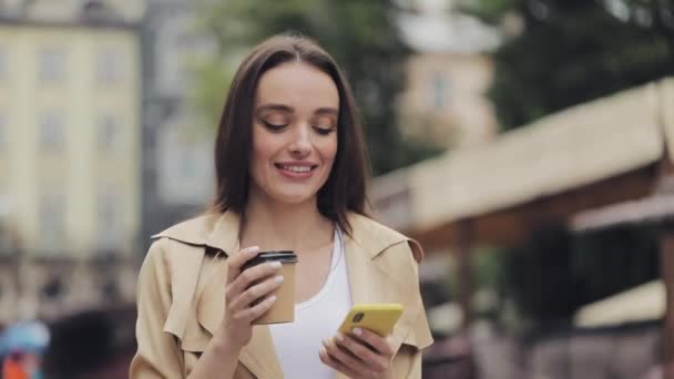 Çekici Genç Kız, elinde kahve fincanı tutarken Smartphone 'unu kullanarak gülümsüyor ve caddede yazı yazarak yürüyor.. — Stok video