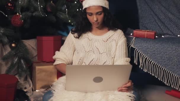 Hermosa joven mulata chica usando sombrero de santas sentado bajo el árbol de Navidad usando su computadora portátil, charlando, haciendo cara sorprendida y diciendo Wow. Concepto de Vacaciones y Año Nuevo . — Vídeo de stock