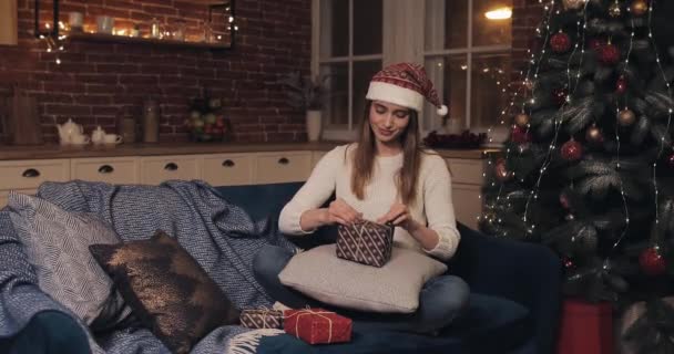 Νεαρό χαρούμενο όμορφο κορίτσι με καπέλο Χριστουγέννων κάθεται στον καναπέ κοντά στο χριστουγεννιάτικο δέντρο προετοιμασία δώρα στο σπίτι έννοια των διακοπών και το νέο έτος. — Αρχείο Βίντεο