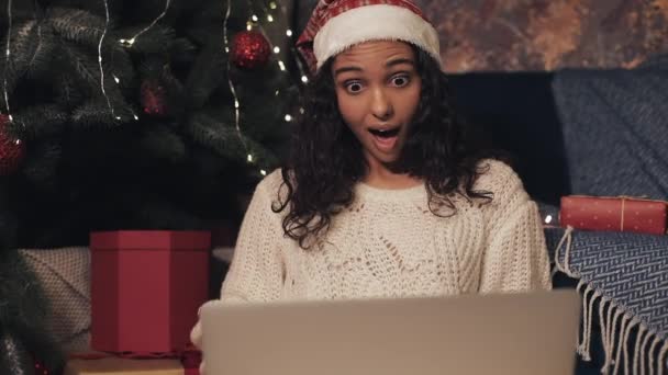 Krásná mladá dívka v klobouku Santas sedí pod vánoční strom pomocí svého notebooku, s videohovoru, dělat překvapený obličej. Koncepce svátků a oslavy nového roku. — Stock video