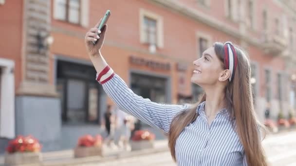 Menina bonita com cabelo castanho e elegante headband vestindo camisa listrada ter uma chamada de vídeo em seu smartphone sorrindo e acenando mão andando na rua da cidade . — Vídeo de Stock