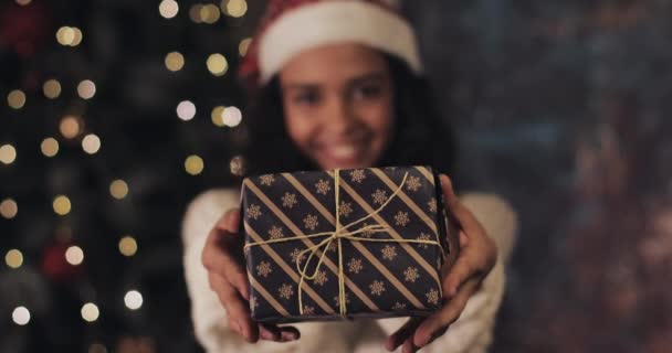 Close Up of Pretty Smiling Mulatto Girl in Santas Hat Gives and Takes Back the Christmas Present Box to Camera, Standing at Decorative Lighting at the Background (en inglés). Concepto de vacaciones de invierno . — Vídeos de Stock