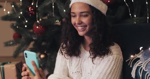 Pretty Young Mulatto Girl in Santas Hatt Sitter under dekorerad julgran och video Chattar med hjälp av sin smartphone, Skrattar, Visar tummen upp, blåser en kyss. — Stockvideo